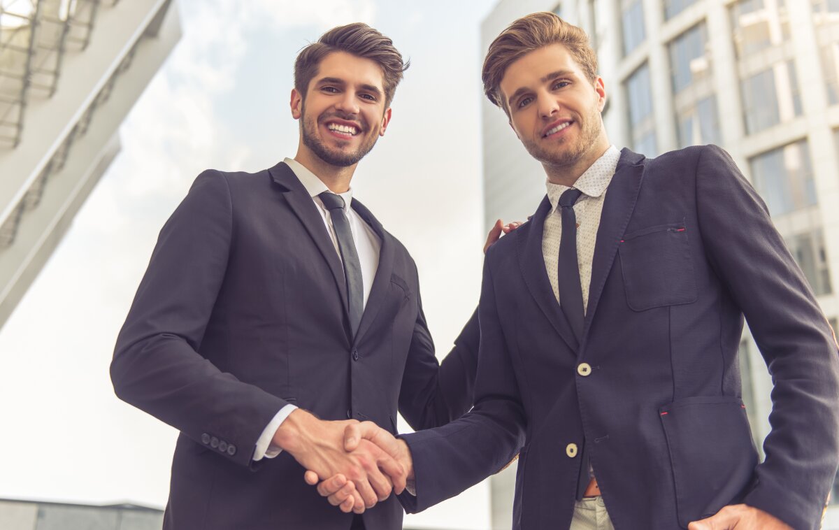 Two business men are shaking hands while looking directly into the camera.